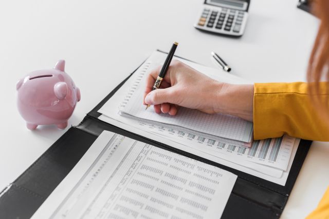 書類にメモをする人と豚の貯金箱・電卓のイメージ